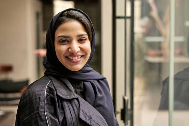 Three-quarter front view of business professional wearing abaya and headscarf pausing from work in modern office to smile at camera.