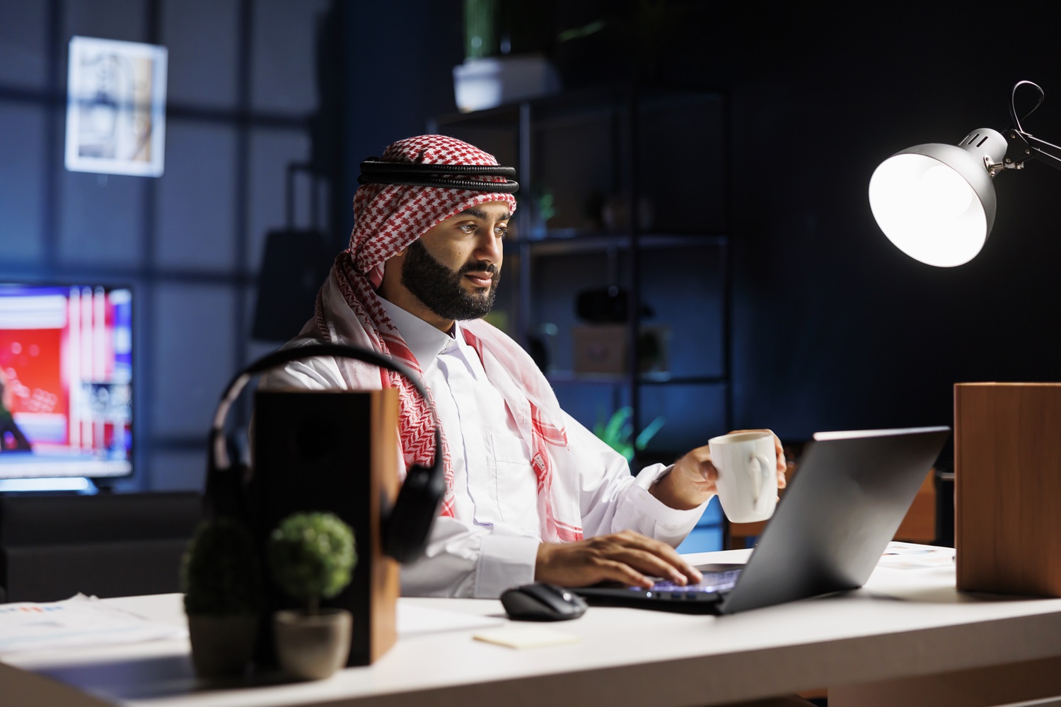 Side-view of a Muslim guy holding a cup of coffee and surfing the net on his digital laptop. Image showcasing a male Arab person engrossed in his personal computer while drinking tea.