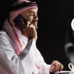 An arab man in traditional attire speaks on a cell phone while browsing on a laptop in an office. Discussing with a person in a phone call on his mobile device the project management consultancy in Saudi Arabia. Side-view, close-up.